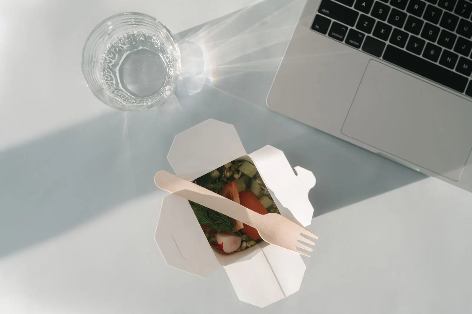 Dizüstü bilgisayarın yanında bir kutu salata ve bir bardak su ile masa başı öğle yemeği